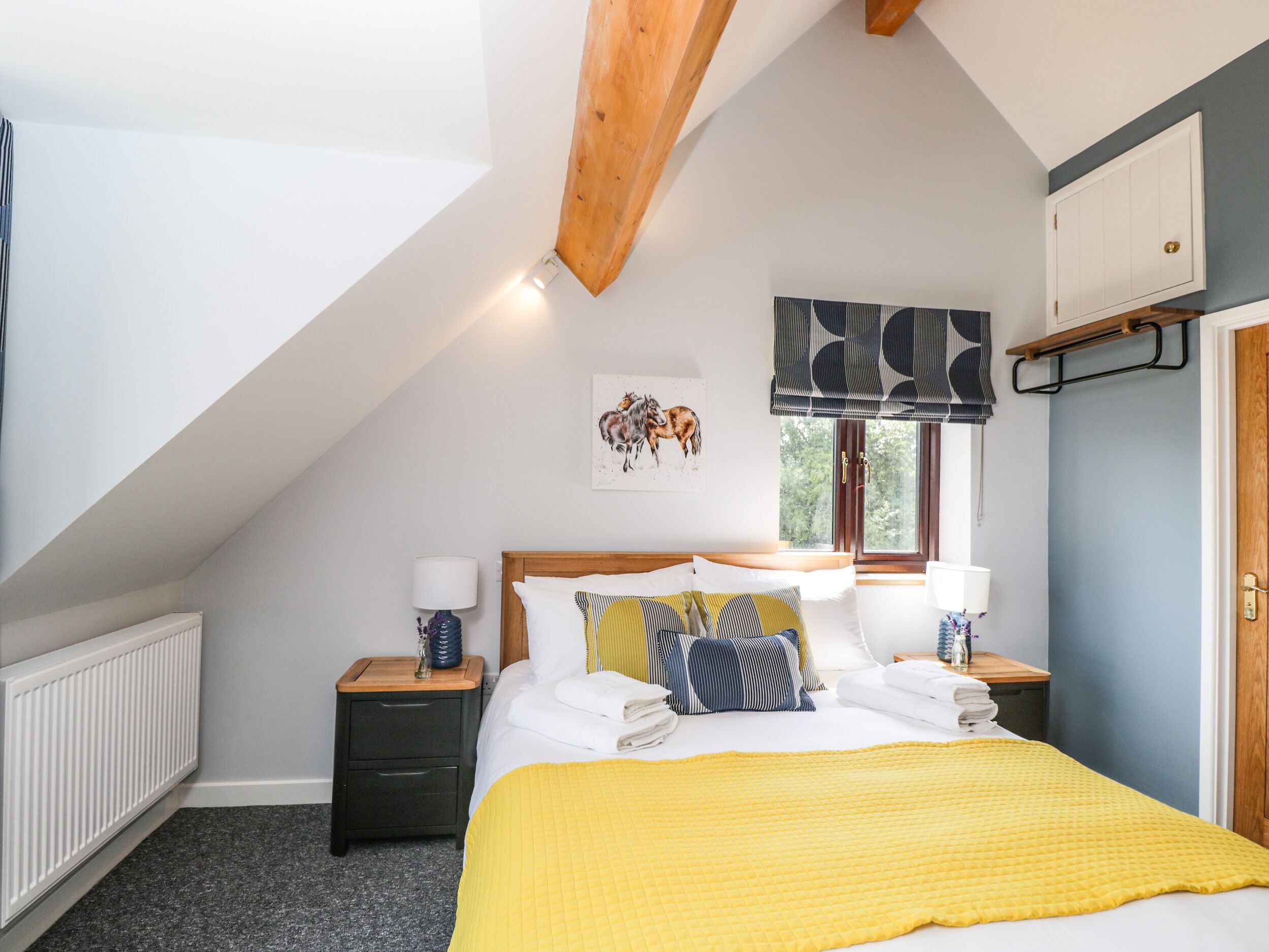 A bedroom with a sloped ceiling, a bed with a yellow quilt, two bedside tables with lamps, a radiator, a window with a view, and a door with a coat rack.