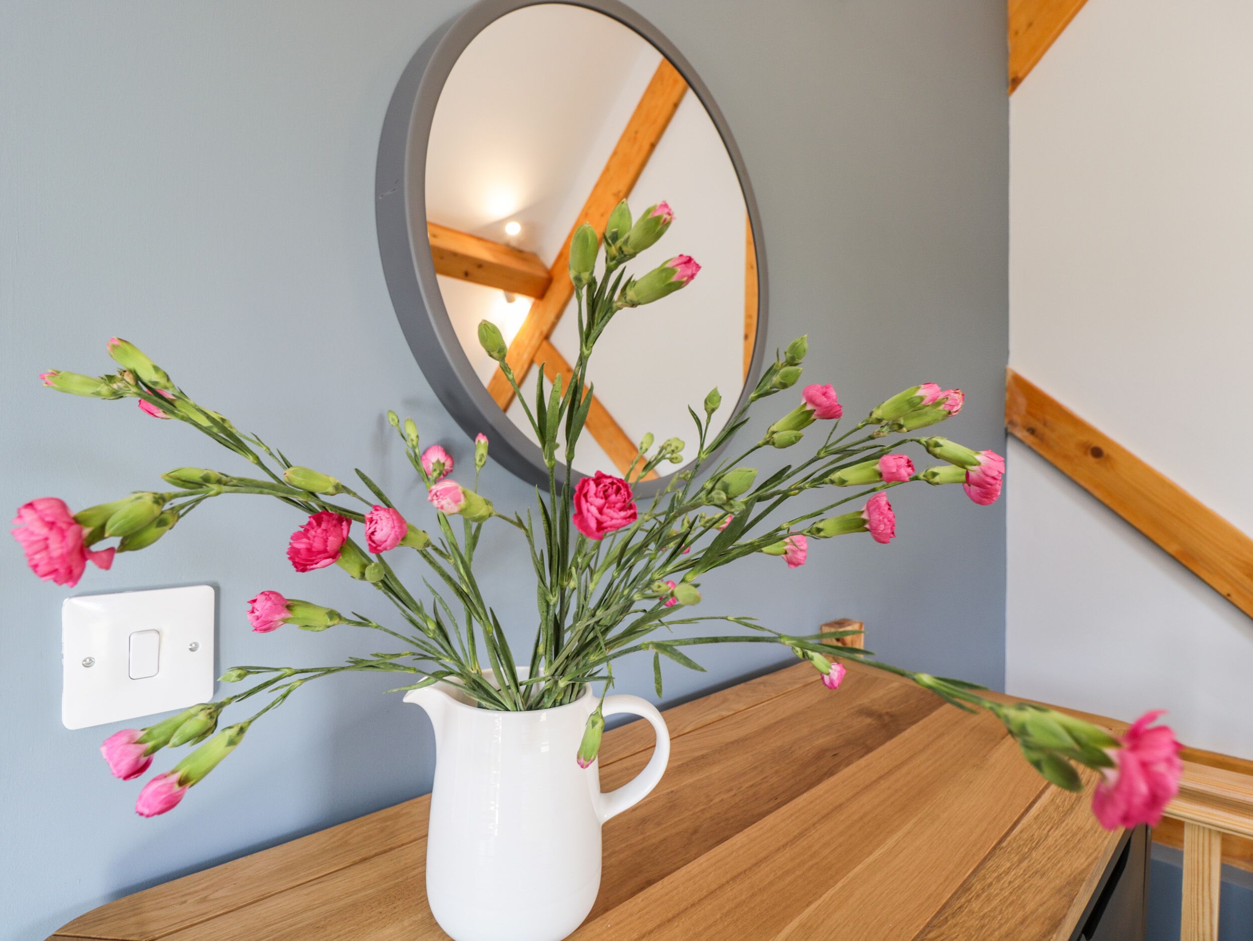 Un pichet blanc orné de fleurs roses, posé sur une surface en bois, un miroir et un interrupteur sur le mur.