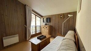 A bedroom with a bed, a desk, a chair, a TV, and a window with curtains.