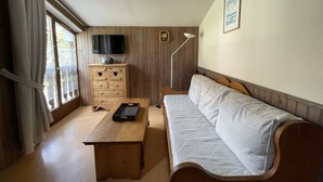 A room with a wooden floor, a wooden desk with a keyboard, a white sofa, a wooden cabinet with drawers, a television on the wall, and a window with curtains.