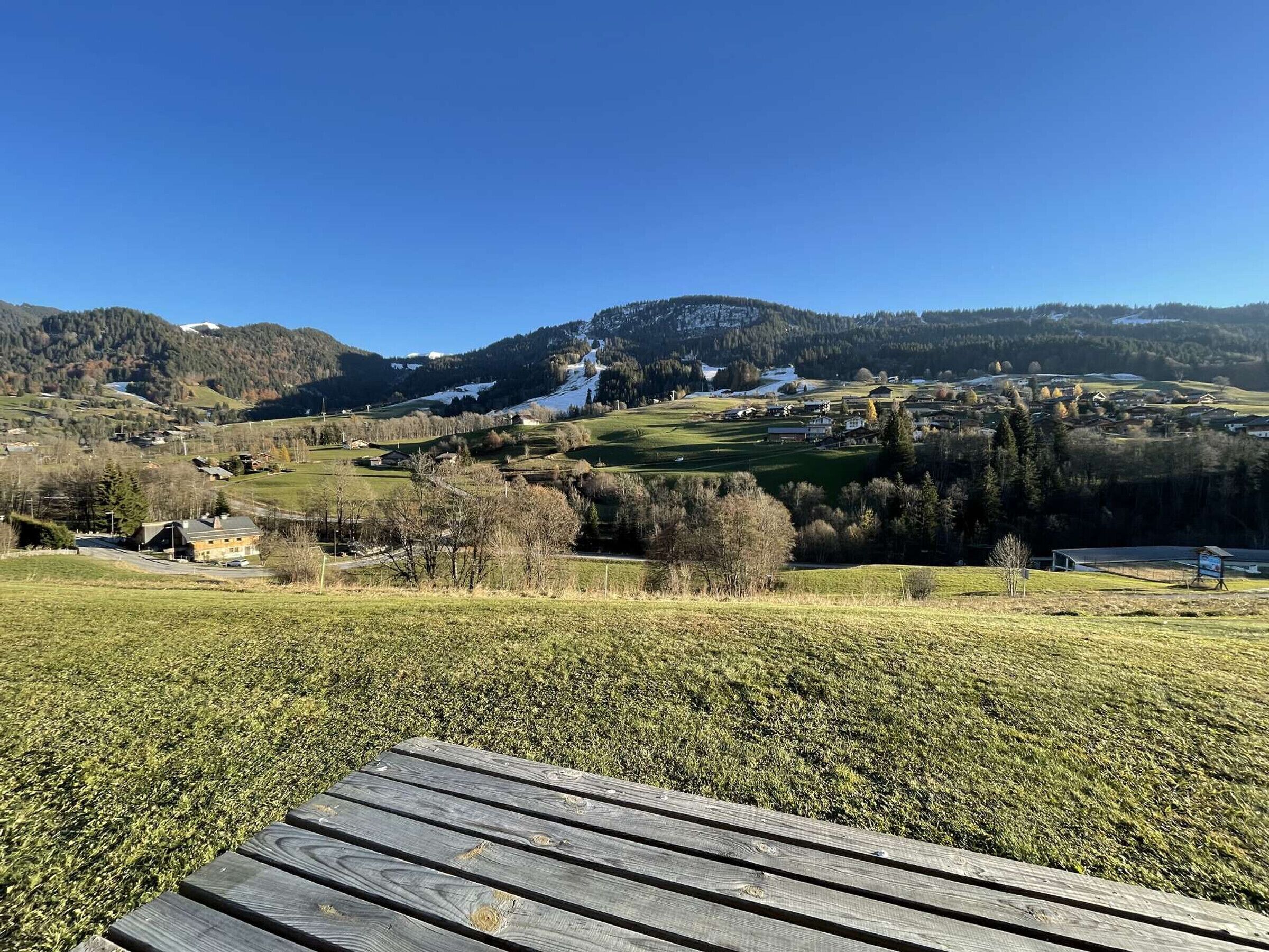 A scenic view of a mountainous area with a village in the valley, a wooden bench in the foreground, and a clear blue sky.