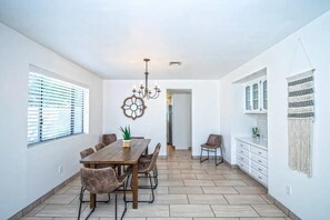 Un vaste espace dédié à la salle à manger, avec une table et des chaises en bois, un lustre et une vue dégagée sur l’extérieur grâce à une porte ouverte.