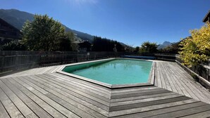 Terraza de madera con piscina, rodeada de valla de madera y árboles, con montañas al fondo.