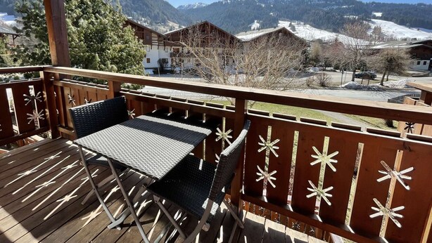 Una terraza de madera con una mesa y sillas, con vistas a un paisaje montañoso con picos nevados y edificios.