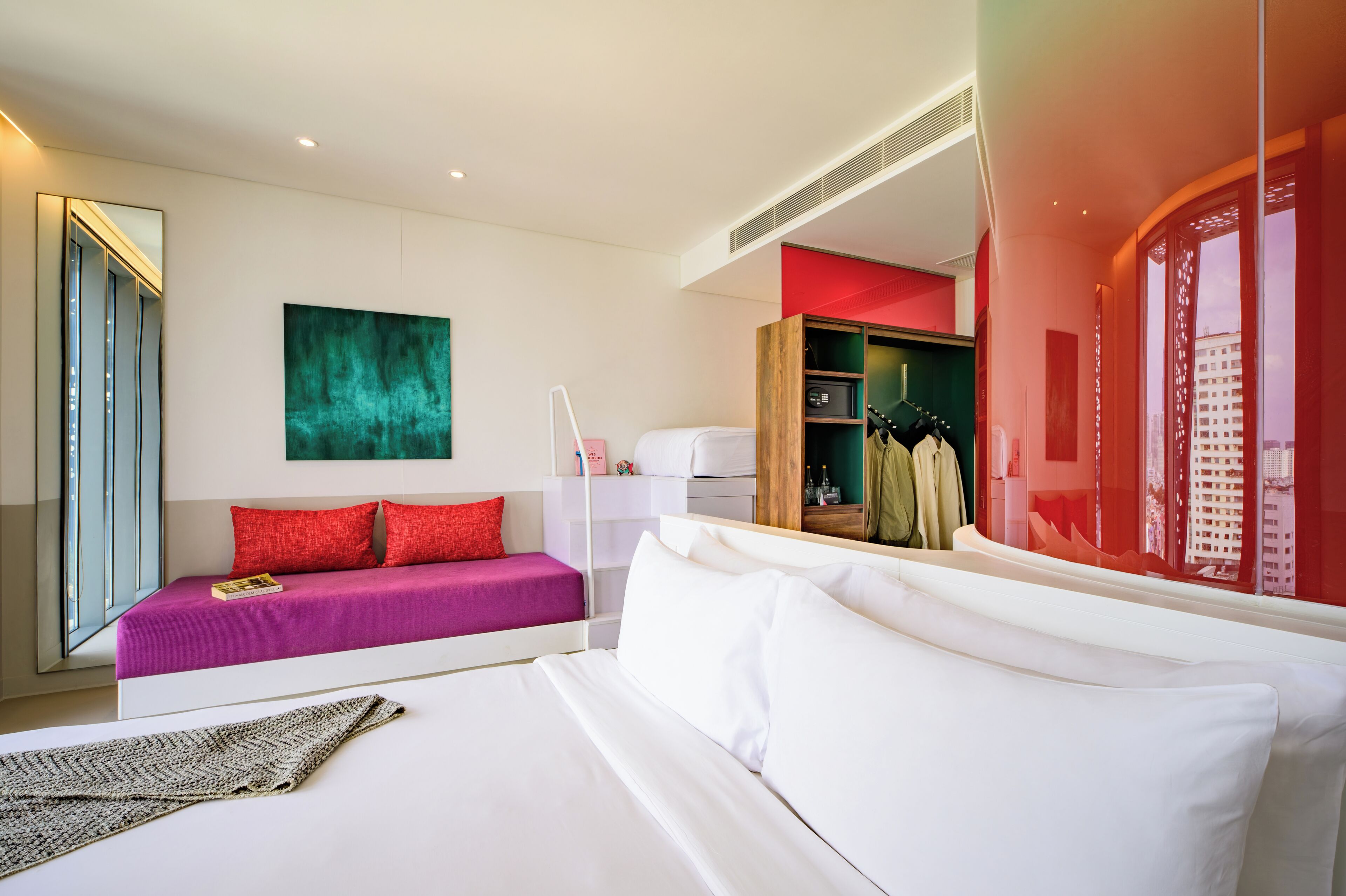 A modern hotel room with a large bed, a sofa, a mirror, and a view of the city through a window.