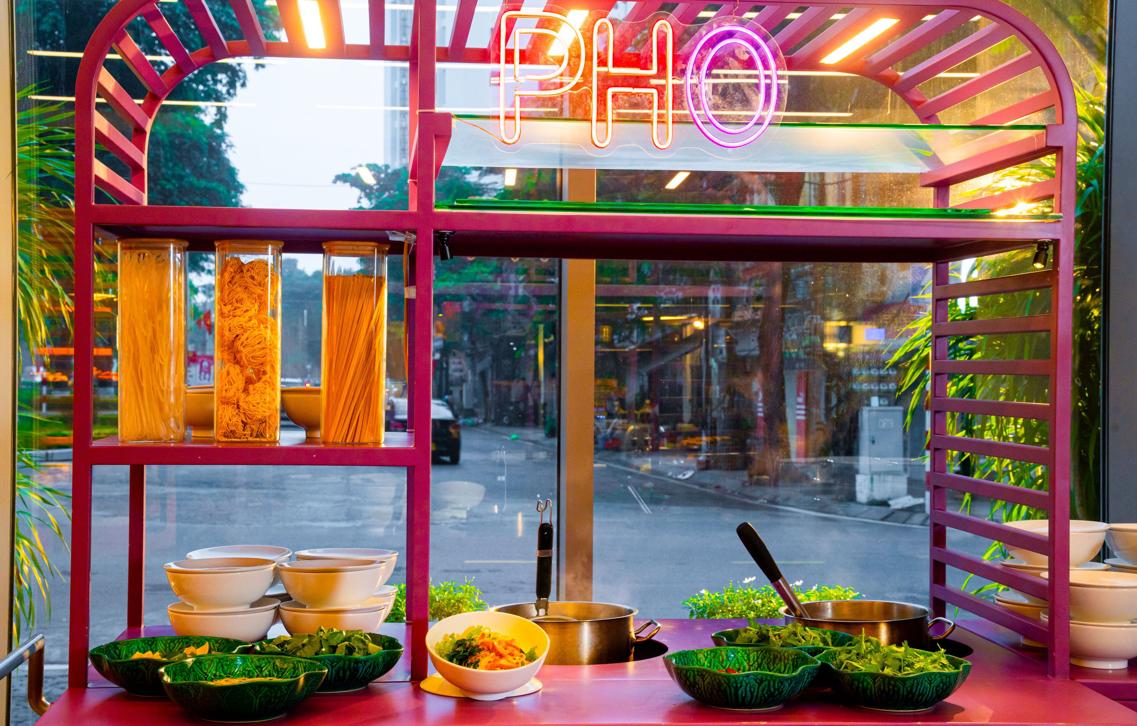 A food stall with a neon sign, glass containers of noodles, bowls, and a pot on the counter.