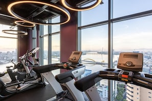 Une salle de sport équipée de matériel d’exercice et dotée de grandes fenêtres offrant une vue sur la ville.