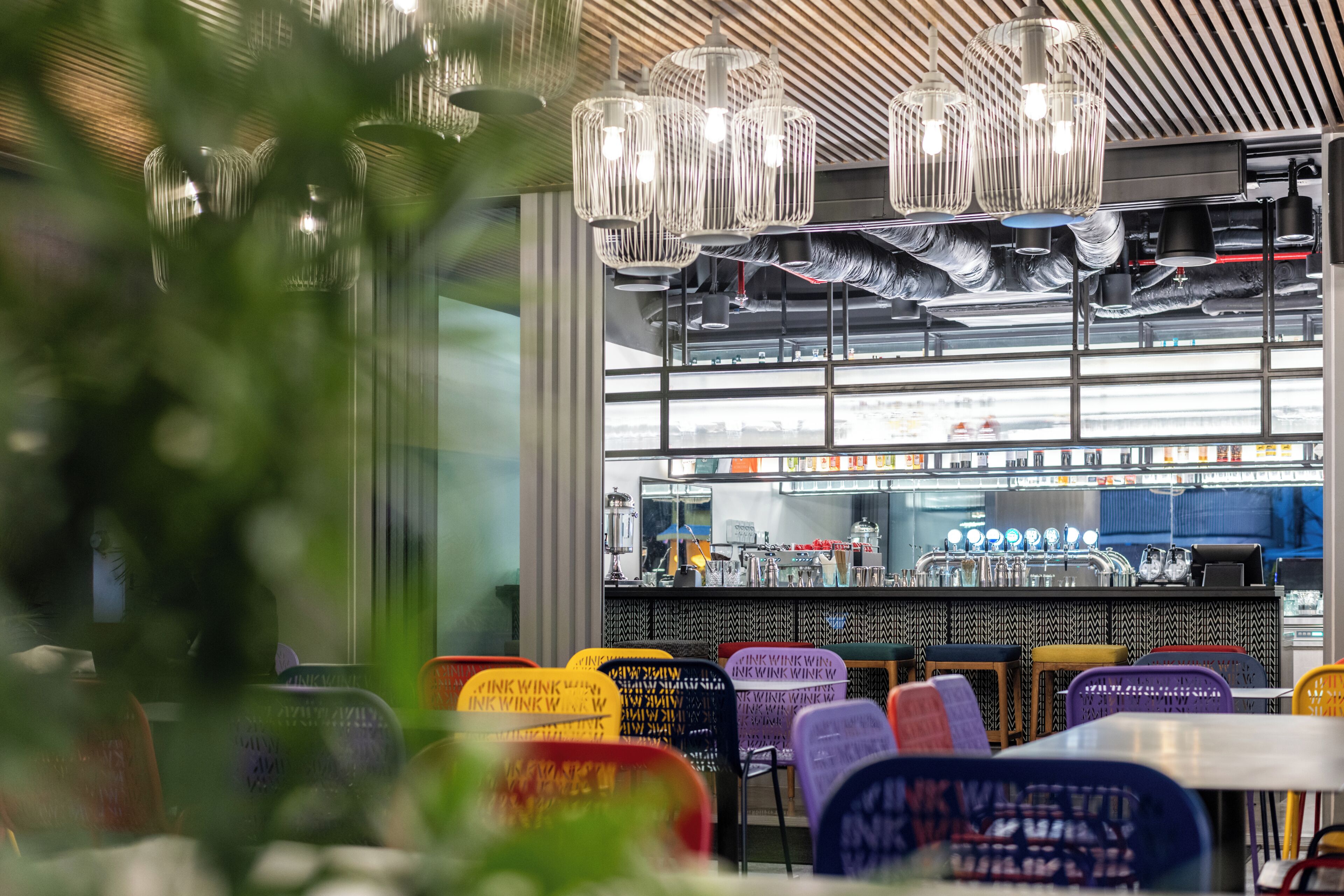 L’intérieur d’un restaurant avec un bar, des tables et des chaises aux couleurs vives, et des lampes suspendues.