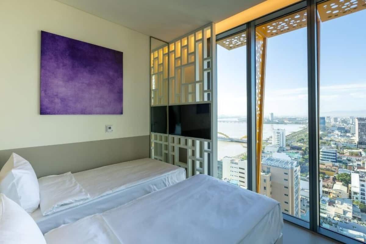 Een hotelkamer met twee bedden, een groot raam met uitzicht op de stad, en een paars kunstwerk aan de muur.