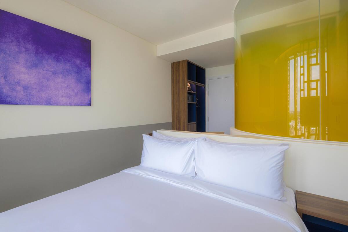 Een hotelkamer met een bed, een paars kunstwerk aan de muur en een groot raam met een gele reflectie.