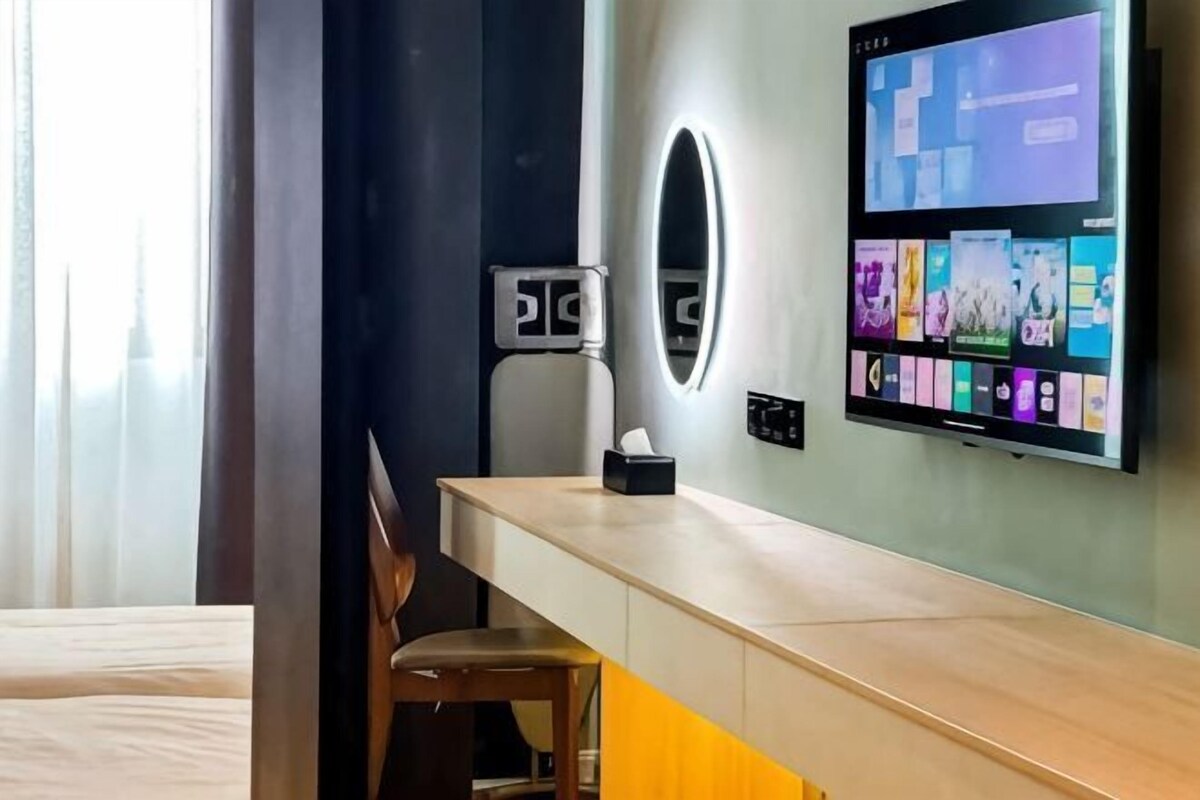 A hotel room with a desk, a chair, a TV mounted on the wall, and a window with curtains.