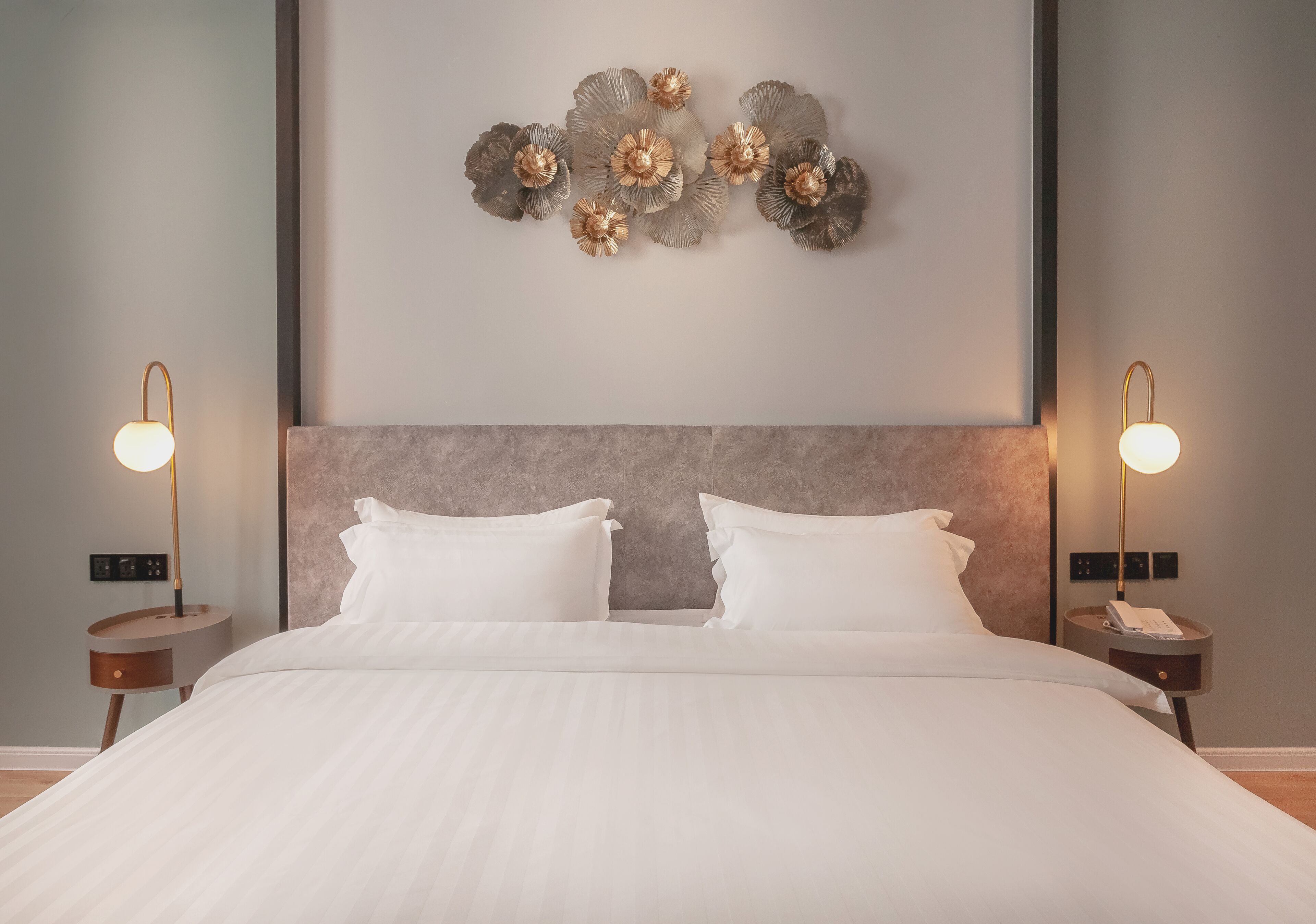 Una cama bien hecha con ropa de cama blanca, un cabecero moderno y un elemento decorativo en la pared sobre ella. Hay dos mesitas de noche con lámparas a cada lado de la cama.