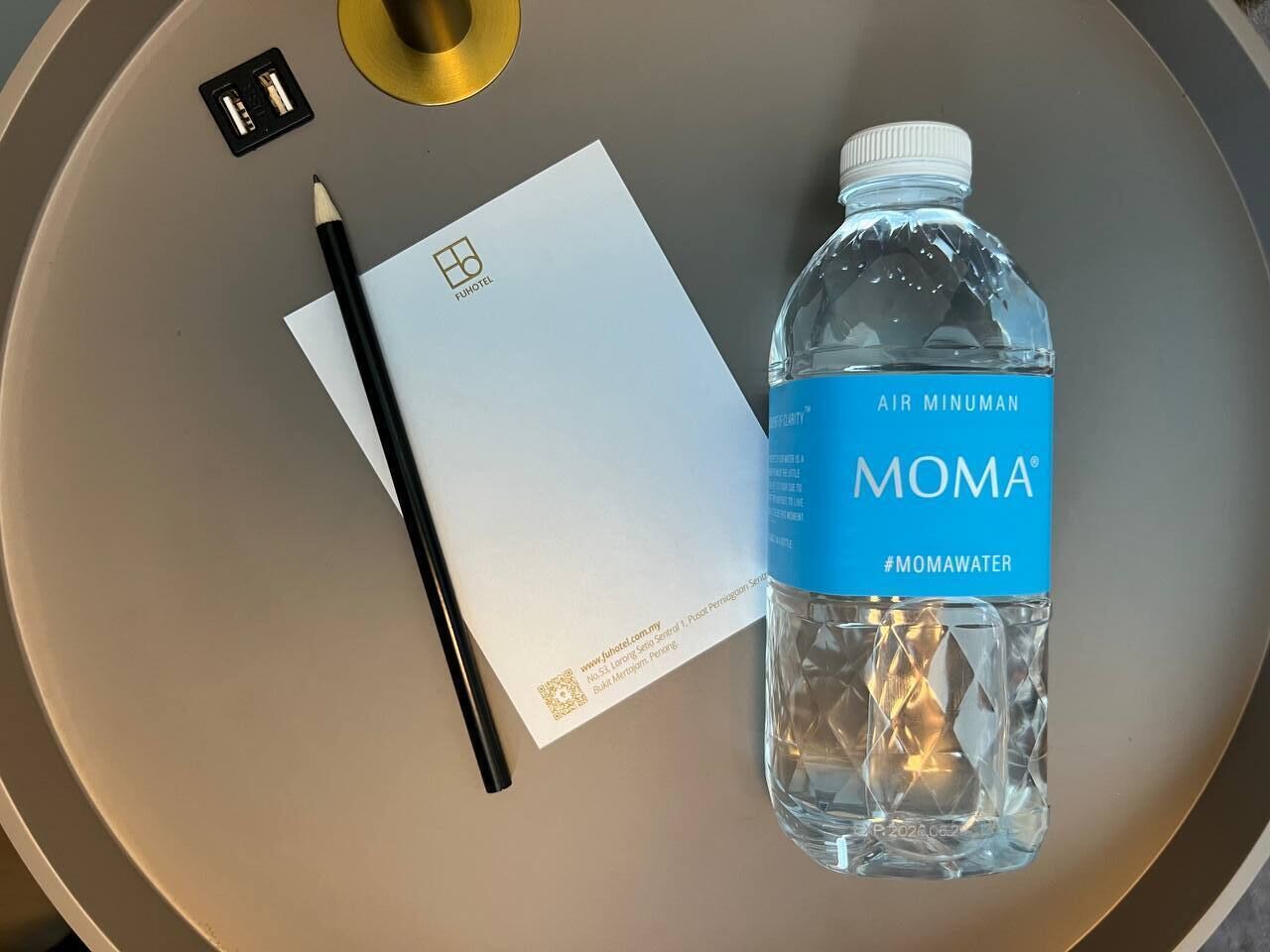 A bottle of MOMA water, a black pencil, a white notepad, and a gold-colored object on a round table.