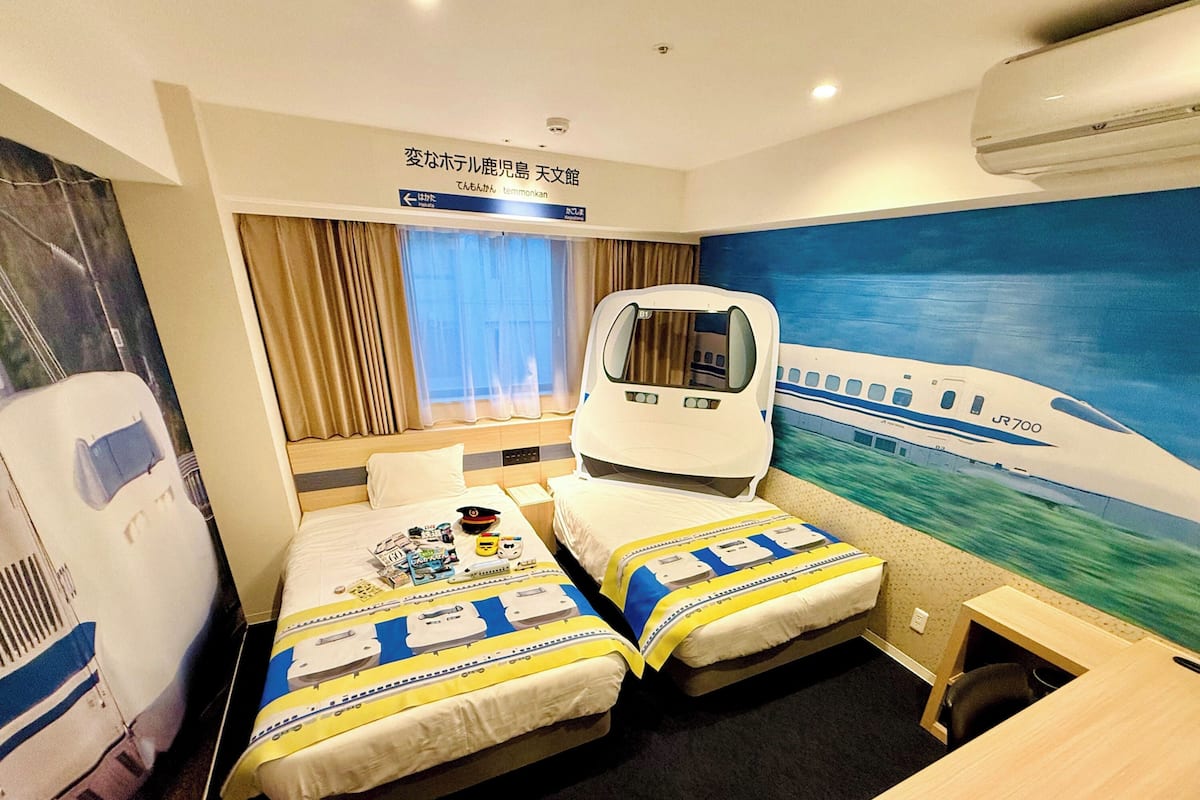 Une chambre d’hôtel avec deux lits, un bureau, une fenêtre avec des rideaux, une fresque murale représentant un train et une inscription en japonais.
