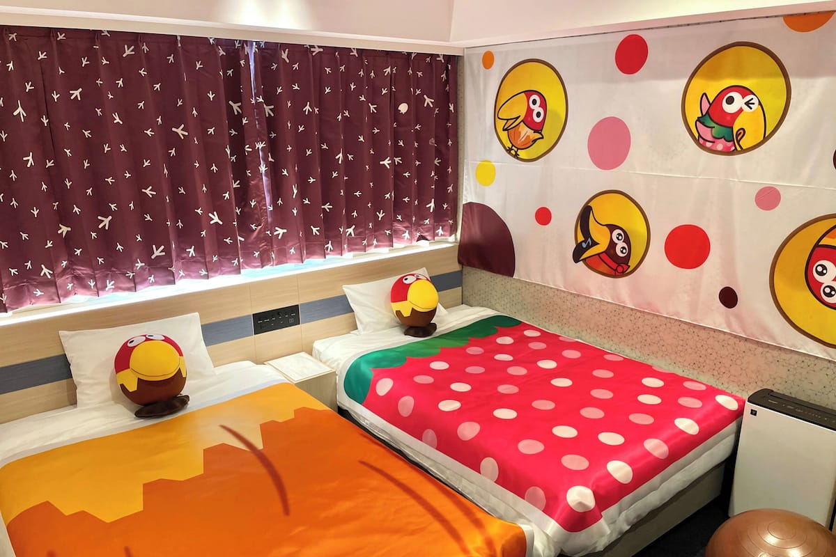 Une chambre d’hôtel avec deux lits, chacun recouvert d’une courtepointe à motifs à pois et d’une autre de couleur unie, une grande fenêtre avec des rideaux et un mur décoré d’autocollants représentant des oiseaux de dessin animé.