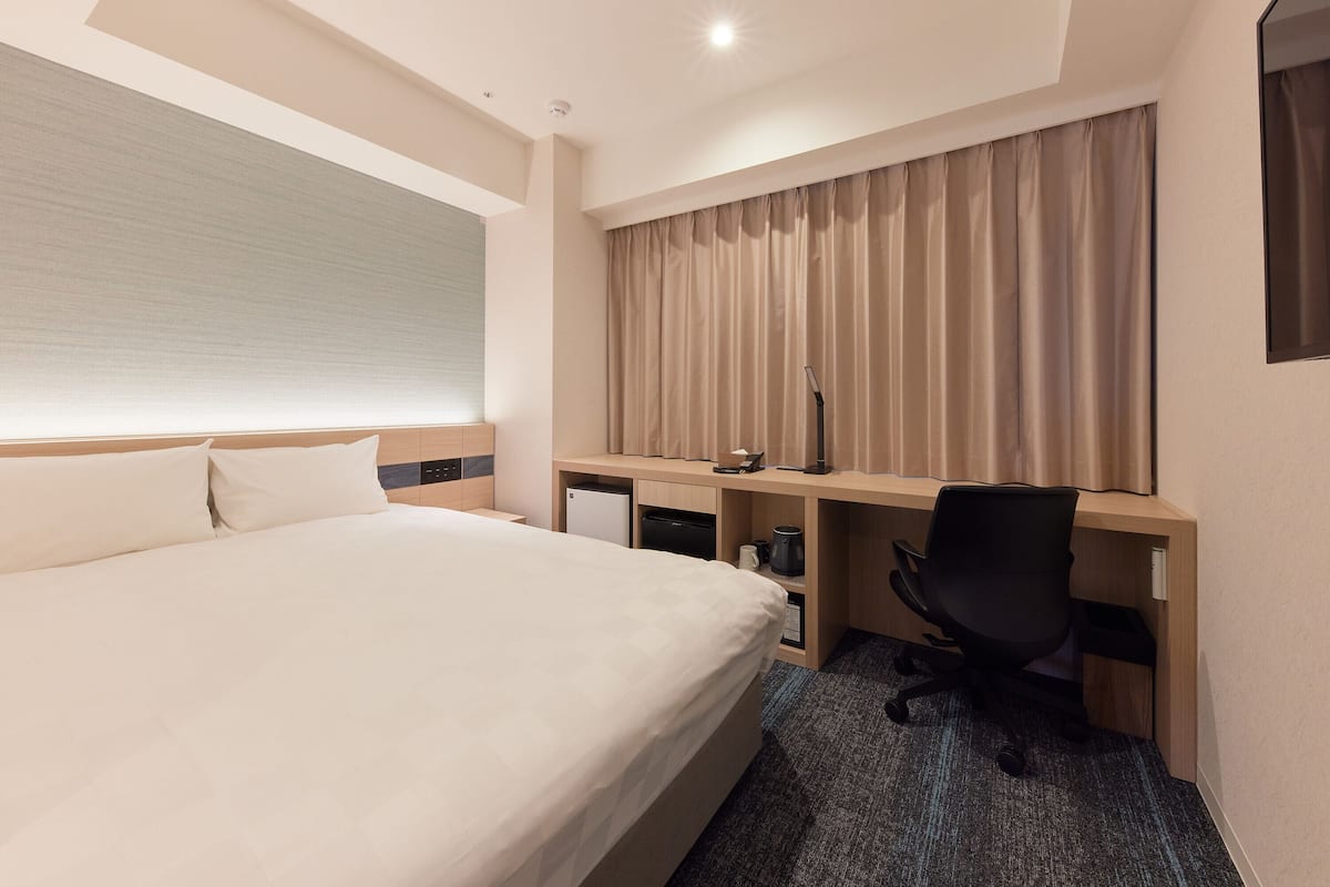 Une chambre d’hôtel avec un grand lit, un bureau avec une chaise, un petit réfrigérateur et une fenêtre avec des rideaux.