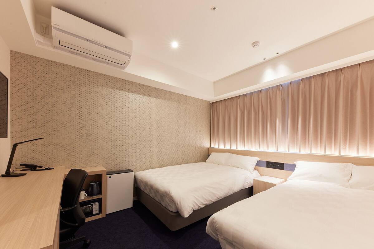 Une chambre d’hôtel avec deux lits, un bureau, une chaise, un petit bureau avec un ordinateur et un appareil de climatisation.