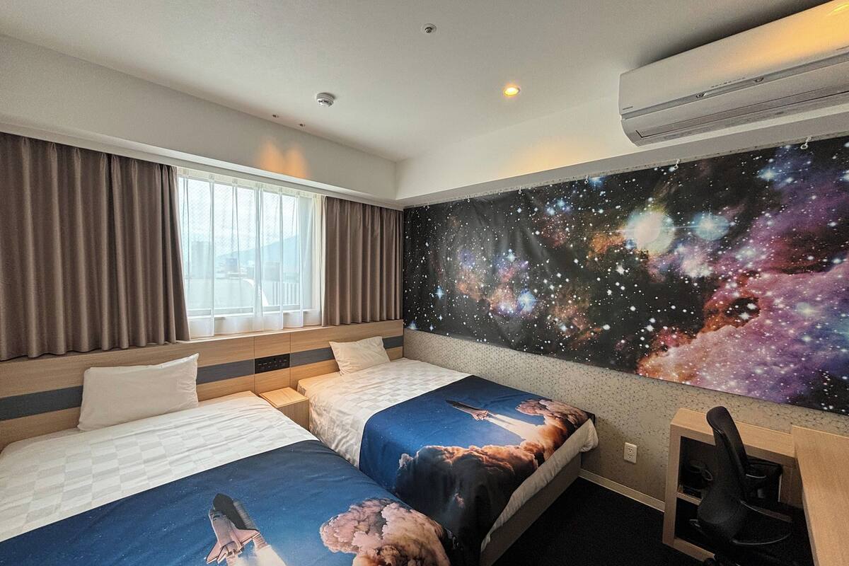 Une chambre d’hôtel avec deux lits, une grande tapisserie sur le thème de l’espace accrochée au mur, un bureau avec une chaise et une fenêtre avec des rideaux.