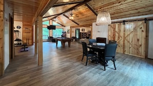 Amplia habitación con suelo de madera y techo alto, que incluye una mesa de comedor con sillas, una mesa de billar y una lámpara colgante.