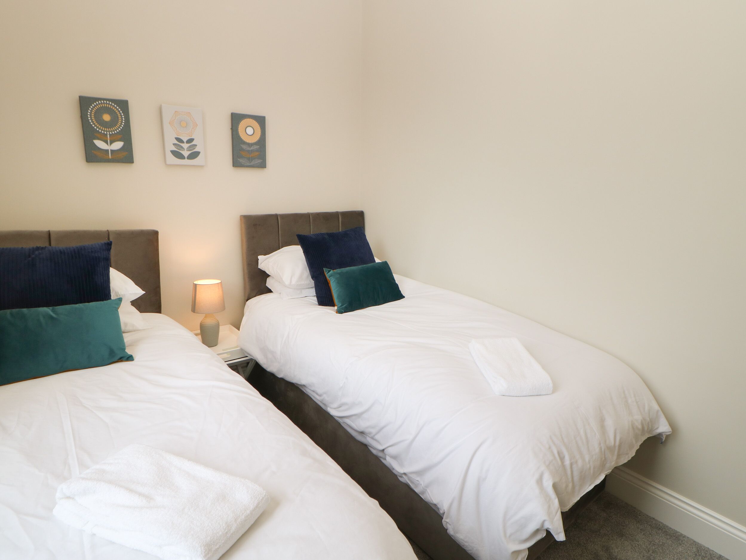A hotel room with two beds, white bedding, teal and blue pillows, a bedside lamp, and three wall art pieces.