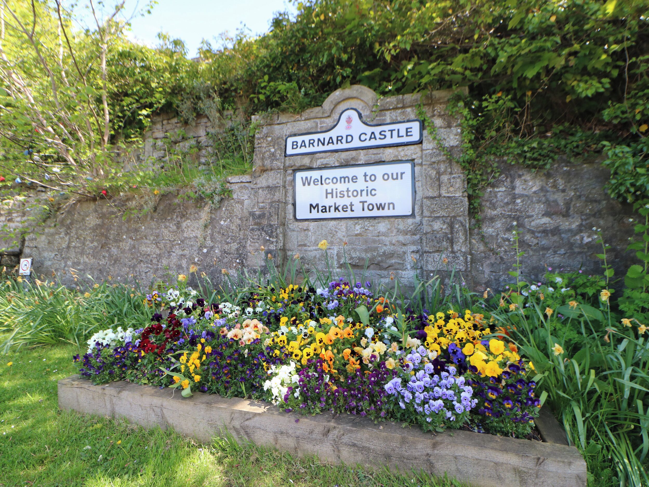 Un panneau accueillant les visiteurs à Barnard Castle, une ville historique réputée pour son marché, avec un mur de pierre et un parterre de fleurs au premier plan.