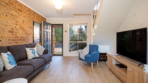 Un salon avec un mur en briques, un canapé, un fauteuil bleu, un téléviseur et un meuble en bois.