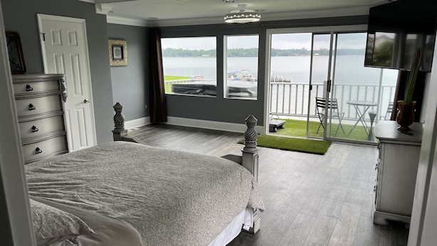 Et soveværelse med en stor seng, et kommode, et fjernsyn og en balkon med udsigt over en sø.