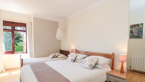 A bedroom with two beds, a wooden headboard, bedside tables with lamps, a window with curtains, and a radiator. - Heselwood (Lyme Regis)
