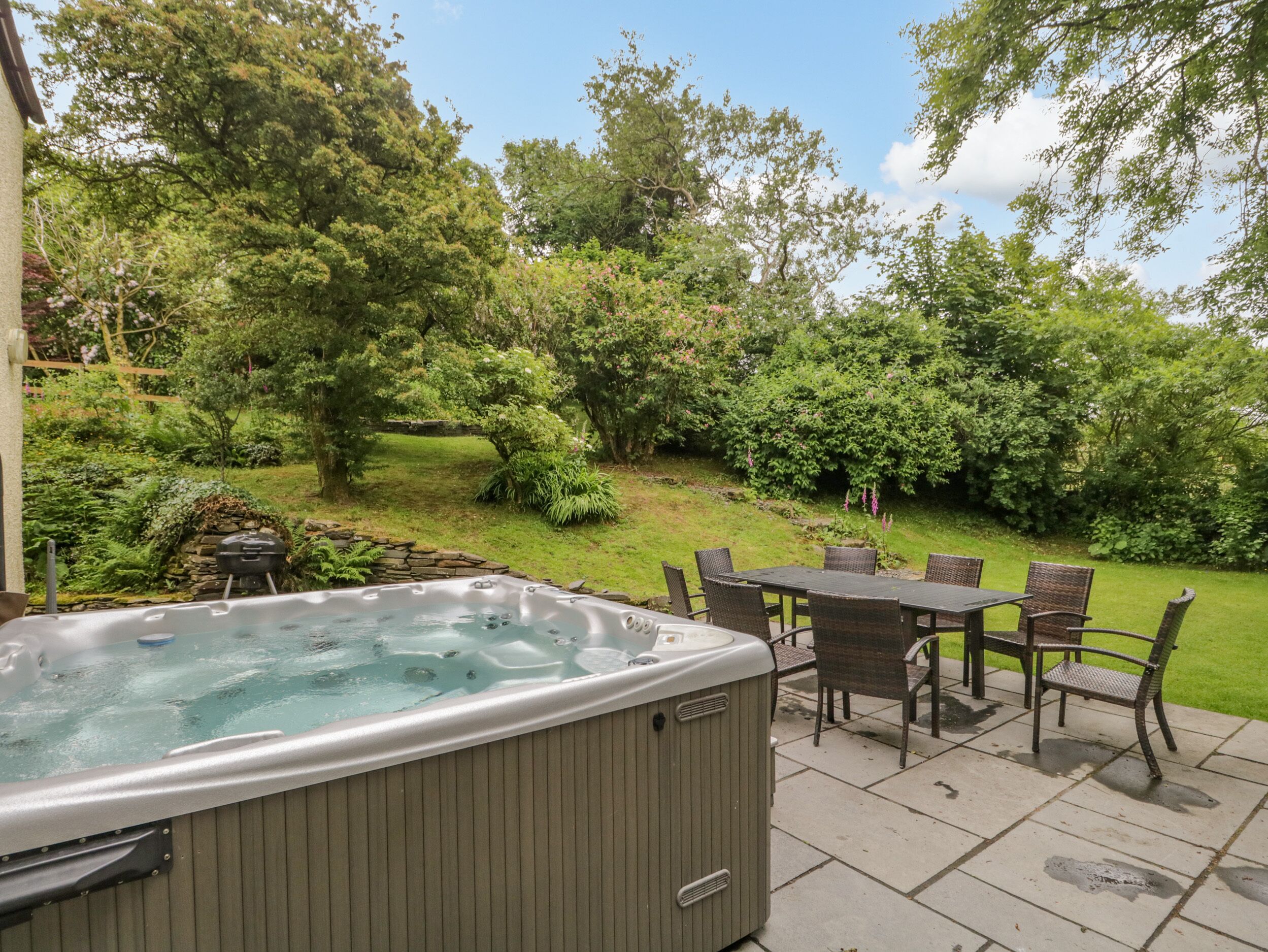 Een hottub met een stenen rand, een tafel met stoelen en een weelderige tuin op de achtergrond.