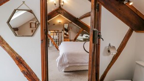 A bedroom with a sloped ceiling, wooden beams, a bed, a toilet, a mirror, and a chandelier. - Ewedale Farm (Ulverston)
