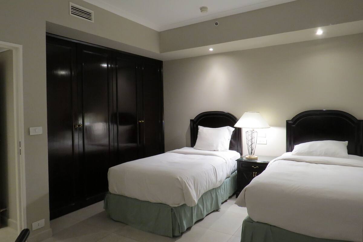 A hotel room with two beds, a dark wardrobe, a bedside table with a lamp, and a ceiling vent.