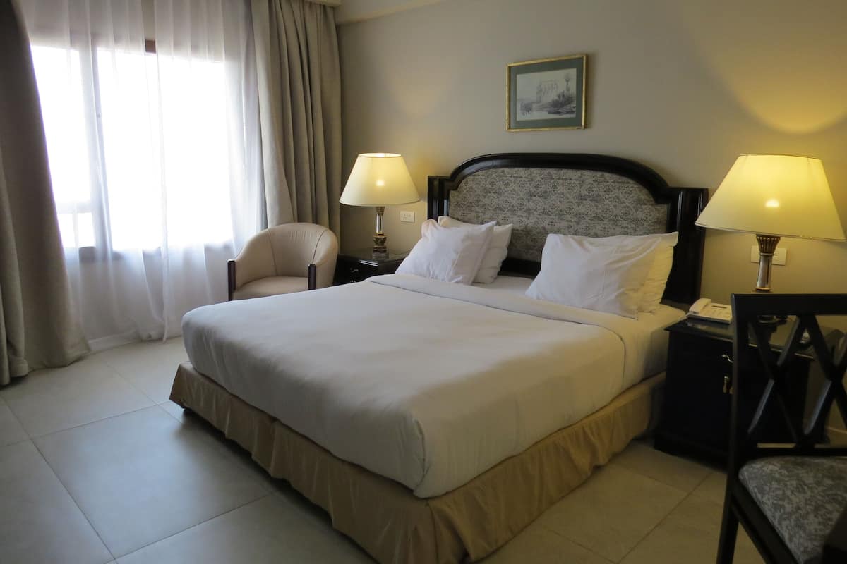 A hotel room with a large bed, two bedside lamps, a chair, a nightstand, and a window with curtains.