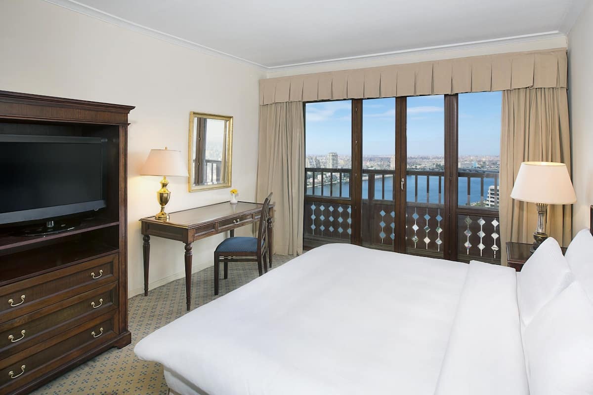 A hotel room with a large bed, a TV, a desk, and a view of a cityscape through large windows.