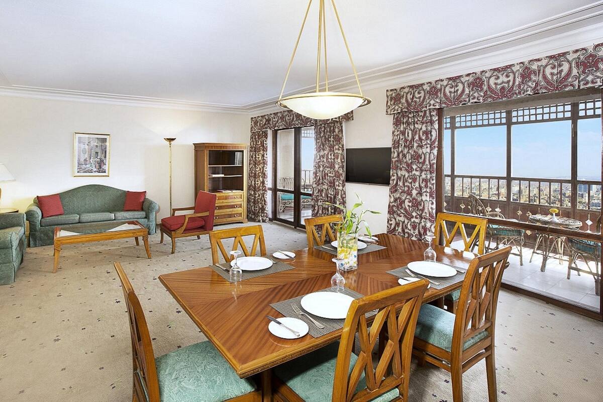 A well-appointed living room with a dining area, a sofa set, a bookshelf, and a large window with a city view.