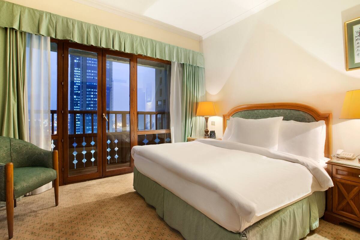 A hotel room with a large bed, a chair, a nightstand with a lamp, and a view of a cityscape through a glass door.