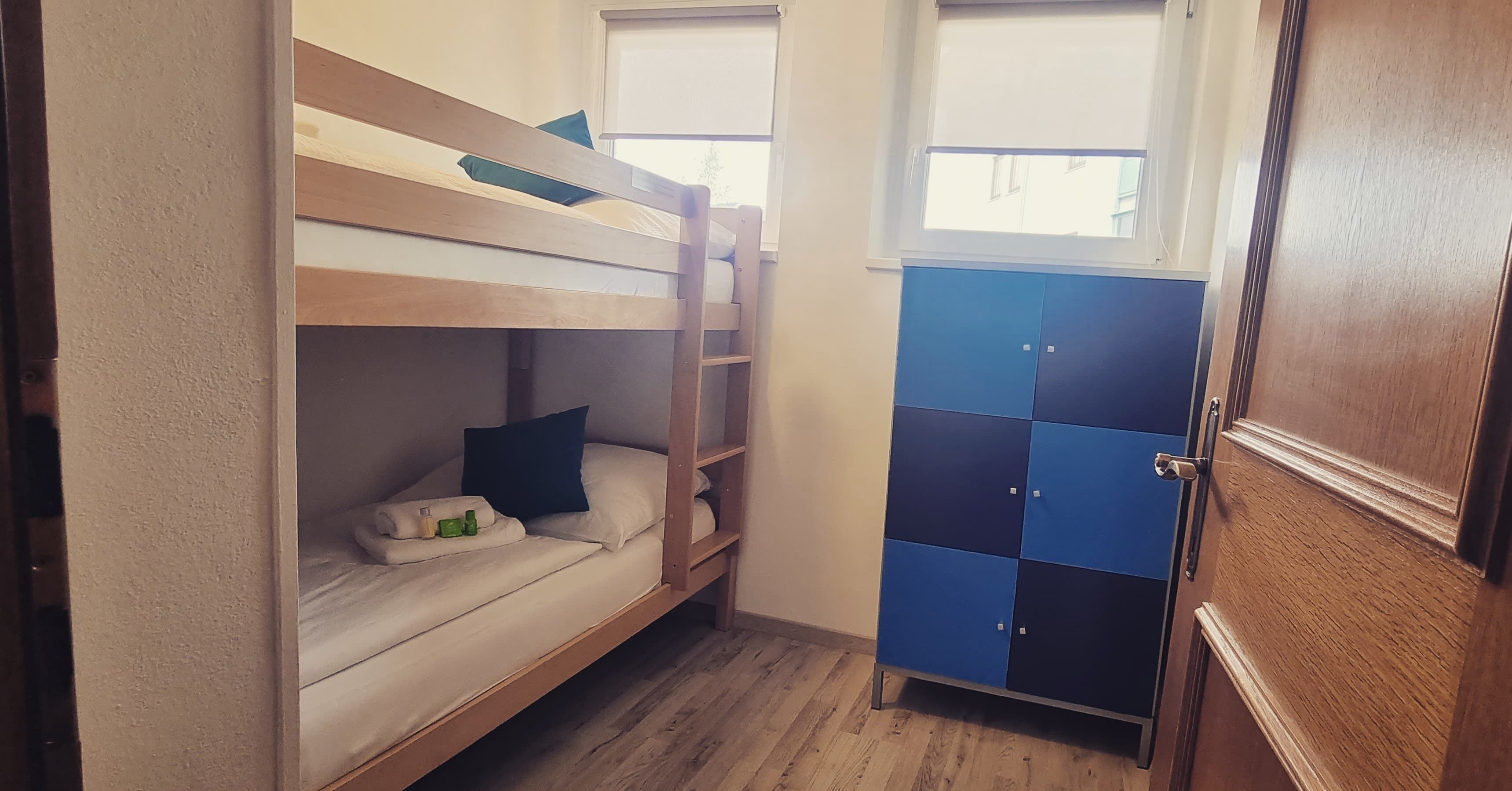 Un letto a castello con una scala in legno, un comodino con una bottiglia e un oggetto verde, e un armadietto blu e nero con una porta.