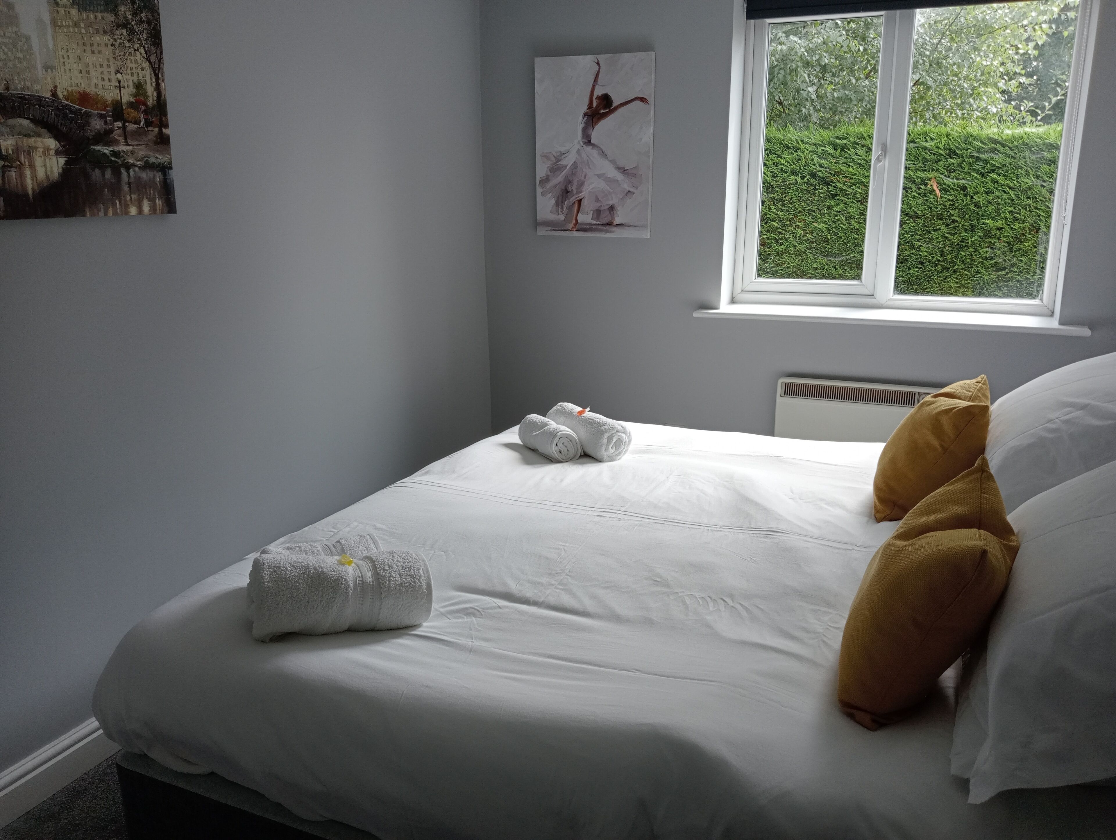 Ein ordentlich bezogenes Bett mit weißen Leinen und zwei Handtüchern, ein Fenster mit Blick auf Grünpflanzen sowie ein Bild an der Wand.