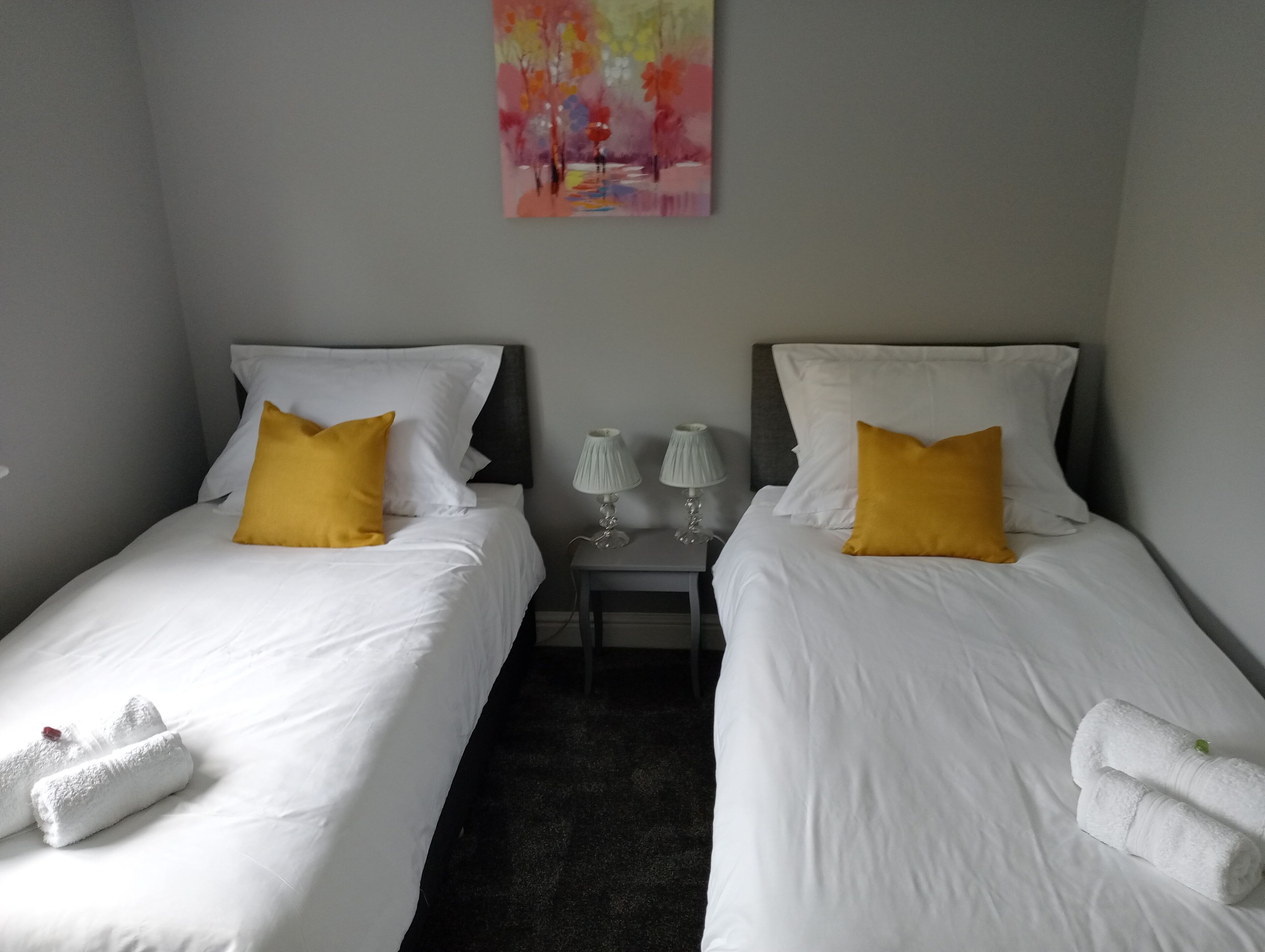 Two single beds with white bedding and yellow pillows, a small bedside table with lamps, and a painting on the wall.