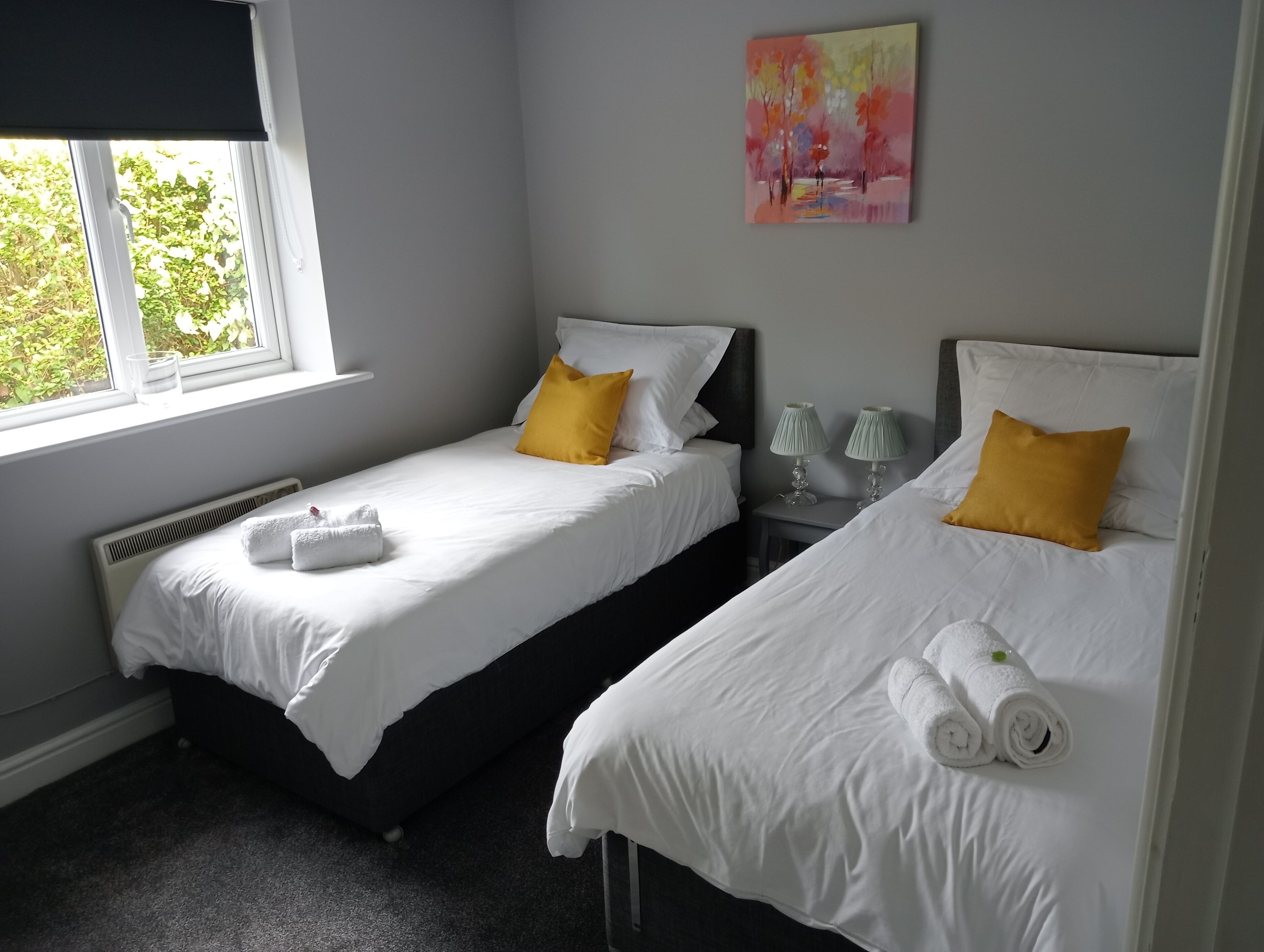 A bedroom with two beds, white bedding, yellow pillows, a painting on the wall, and a window with a view of greenery.