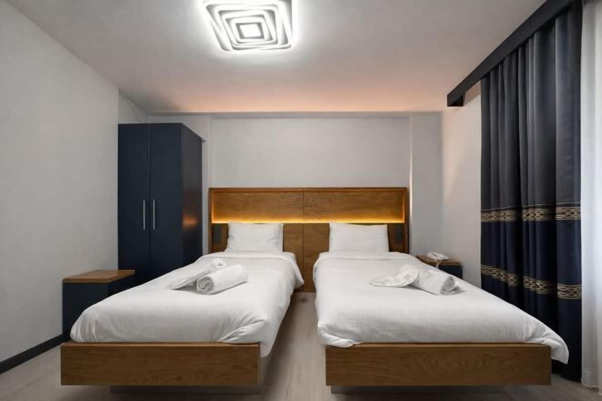 A hotel room with two beds, a wardrobe, a bedside table, and a modern ceiling light fixture.