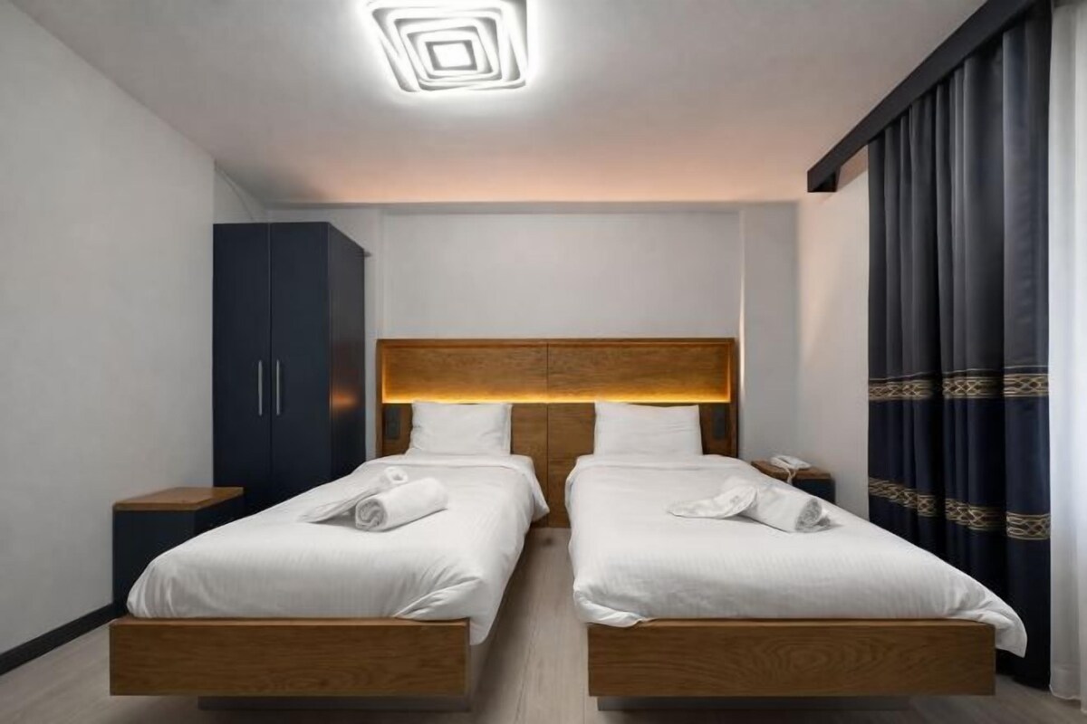A hotel room with two beds, a wardrobe, a bedside table, and a modern ceiling light fixture.