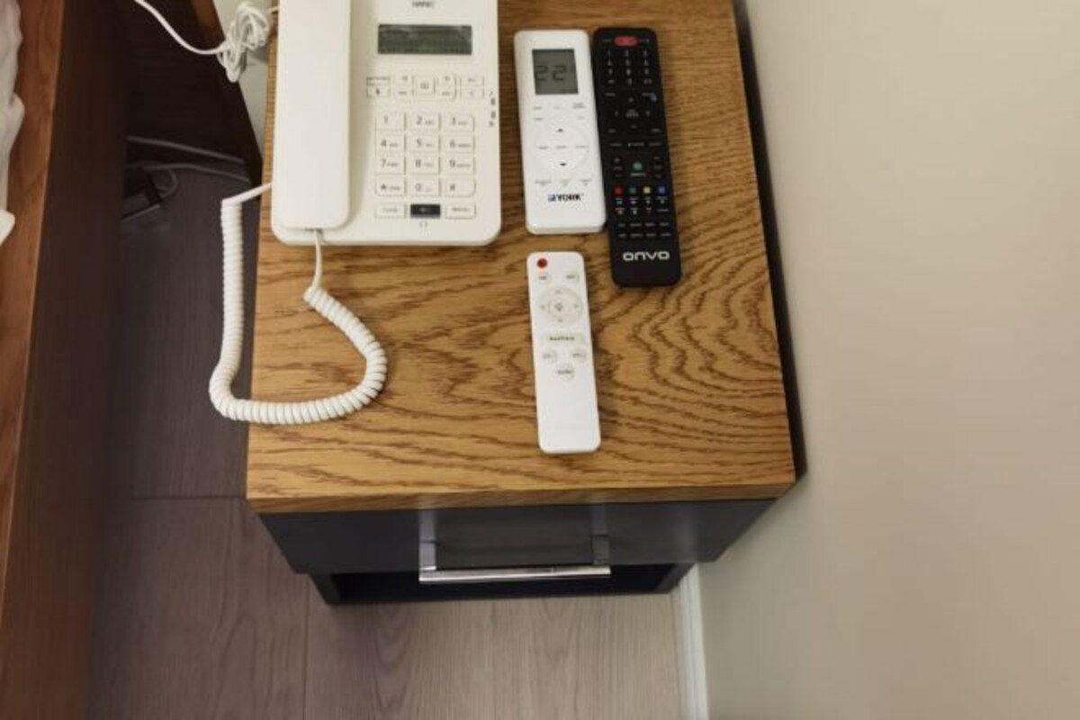A wooden desk with a white telephone, a digital clock, and a remote control.