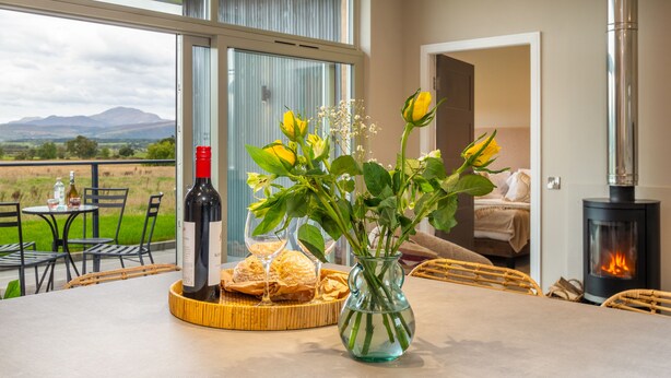 Un coin repas avec une table dressée pour deux personnes, une bouteille de vin, du pain et un vase rempli de fleurs.