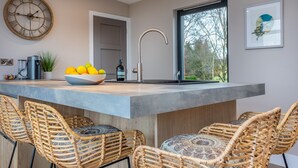Une cuisine moderne dotée d’un grand îlot central, de chaises en osier, d’une horloge et d’une fenêtre donnant sur la forêt.
