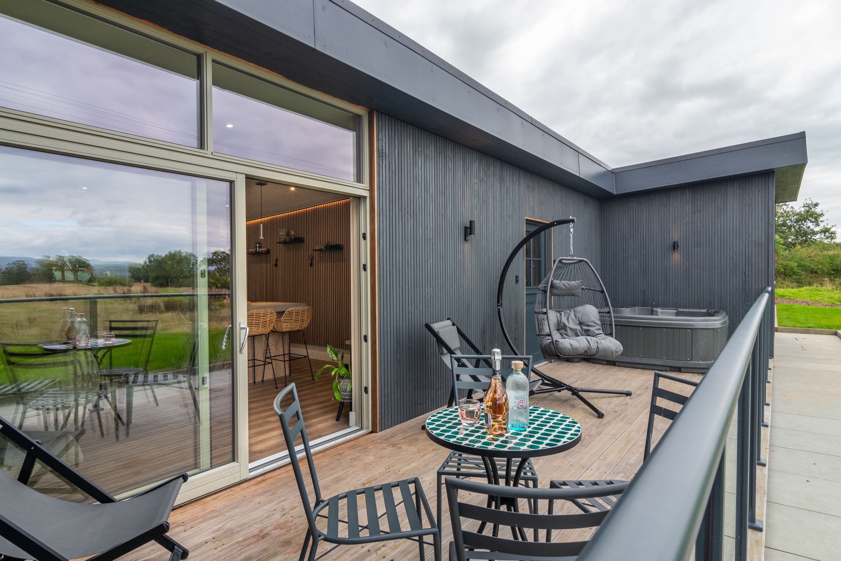 Une terrasse extérieure moderne, dotée d’une porte vitrée donnant accès à un espace repas, à un jacuzzi et à un coin salon avec une table et des chaises.