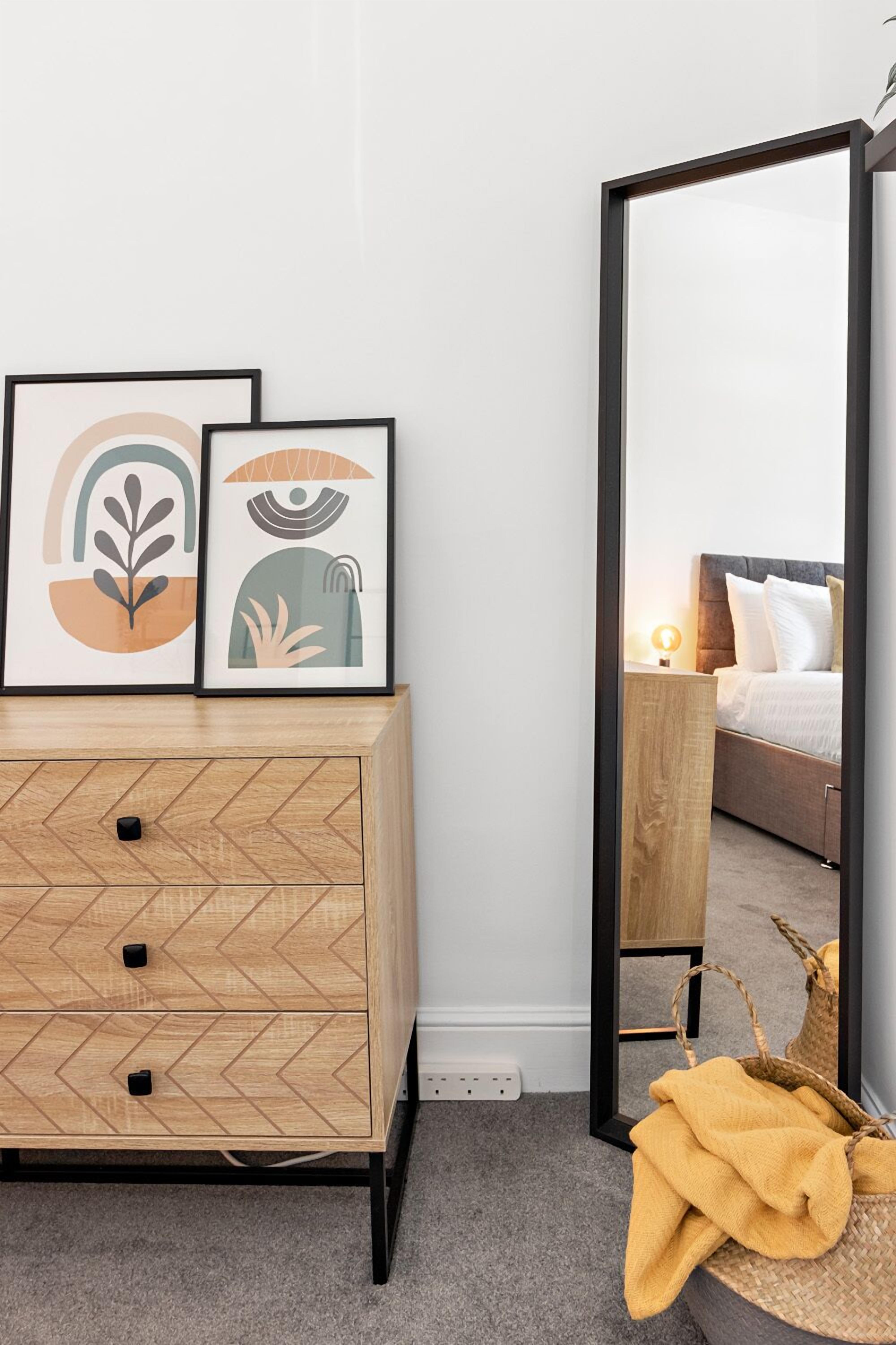 A bedroom with a bed, a wooden dresser with drawers, a mirror, and two framed artworks on the dresser.