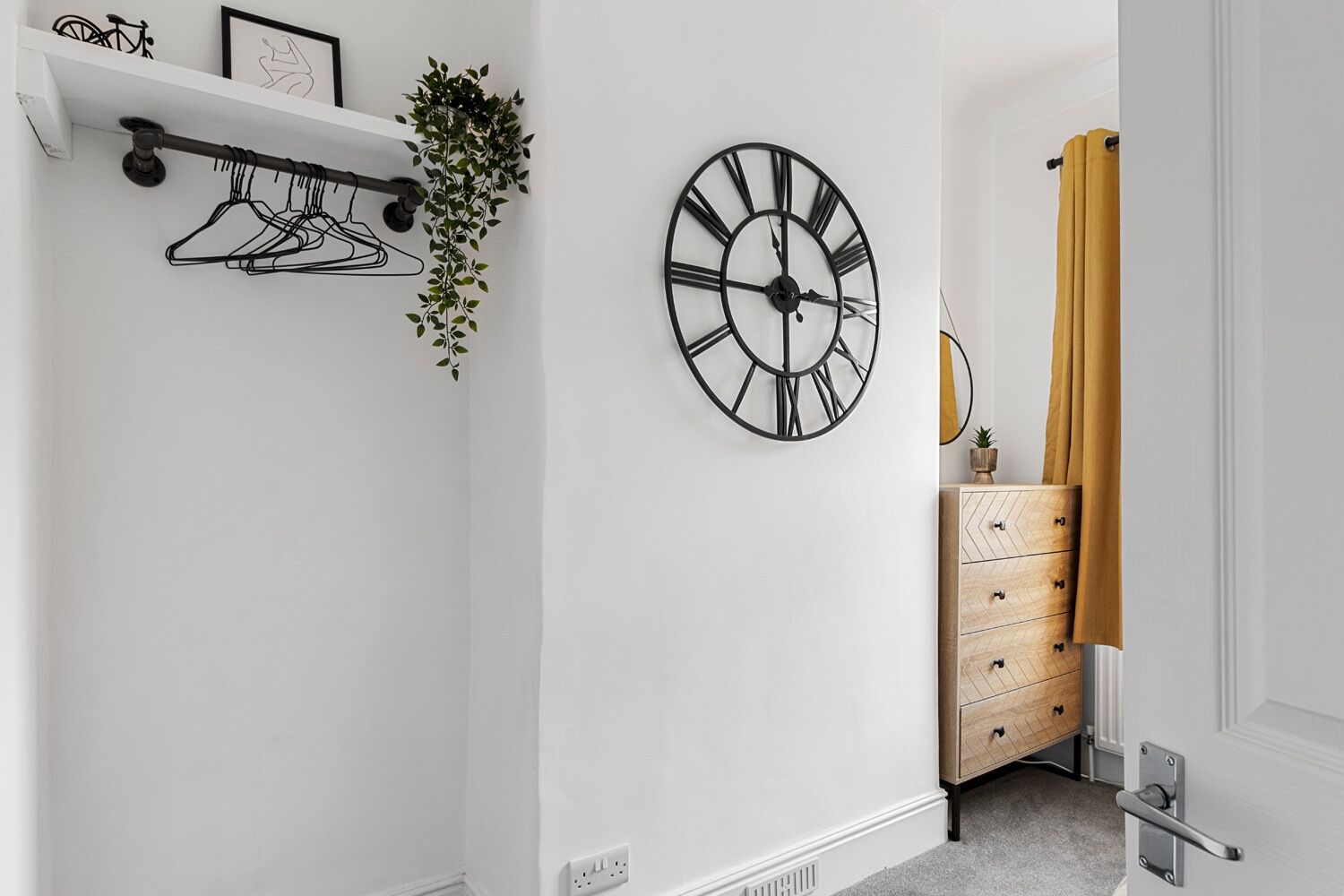 A bedroom with a white wall, a black clock, a wooden dresser, and a yellow curtain.