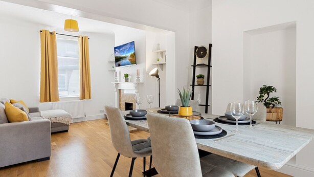 A modern living room with a dining area, featuring a sofa, dining table, chairs, and a TV mounted on the wall. - Church View (Exeter)