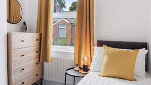 A bedroom with a bed, a dresser, a bedside table with a lamp, and a view of a brick building through the window. - Church View (Exeter)