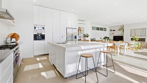 Uma cozinha moderna com armários brancos, uma ilha central com banquetas e uma área de jantar com uma janela ampla.